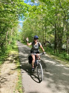 photo taken of a female cyclist in a tree lined pathway, smiling and waving at the camera