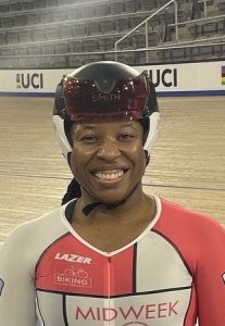 Woman smiling in facing the camera from the shoulders up wearing a time trial cycling helmet with the velodrome track in the background